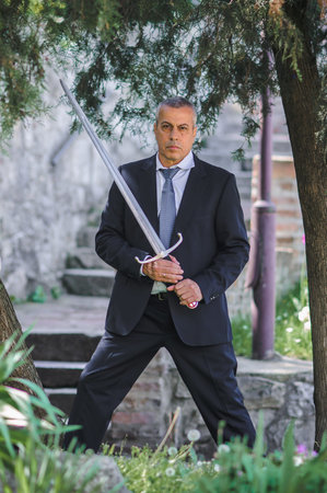 Portrait of handsome businessman model man in a modern casual suit with a large medieval sword....