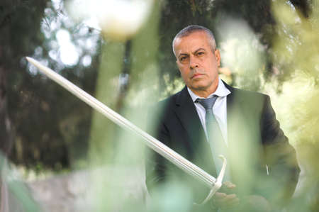 Portrait of handsome businessman model man in a modern casual suit with a large medieval sword. Modern fashion street style concept. Elegant urban warrior. Corporate and money securityの写真素材