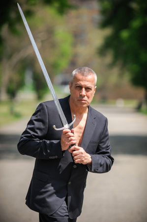 Portrait of handsome businessman model man in a modern casual suit with a large medieval sword....