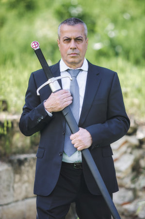 Portrait of handsome businessman model man in a modern casual suit with a large medieval sword....