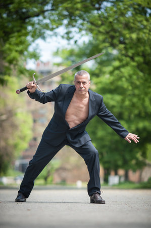 Portrait of handsome businessman model man in a modern casual suit with a large medieval sword....