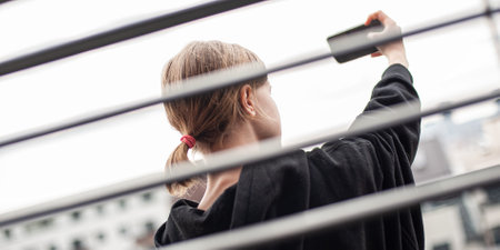 Influencer teenage girl in the city behind open horizontal aluminum metal black blinds, using a smartphone. Close-up, window background. Back view, bright, texture.の写真素材