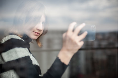 Influencer teenage girl in the city by the window, using a smartphone. Background reflection of the sky on the window. Side view, bright, texture.の写真素材