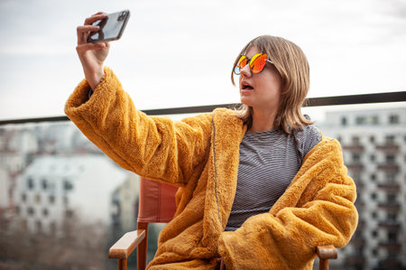 Beautiful young girl in a yellow fur coat and red sunglasses is sitting in a director's chair on the terrace of a modern building. Use smartphone, social media influencer, taking picture, selfie, posing, city in background.の写真素材