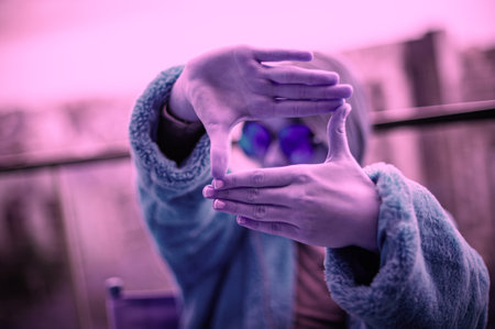 Capture Moments. Young girl in a fur coat and blue glasses creates a frame with hands, city in background.の写真素材