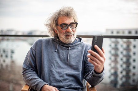Handsome senior man with grey hair and eyeglasses sitting on terrace, using cigarette and smartphone, communicating, having fun and enjoyingの写真素材