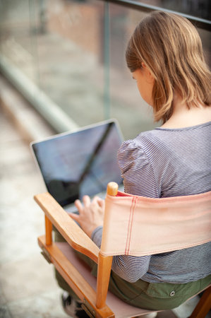 Pretty teenage girl social media influencer is sitting in the director's chair and using a laptop while on the balconyの写真素材
