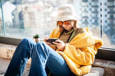 Copy space. Beautiful teenage girl, internet influencer sitting on patio floor on balcony, using smartphone, taking a selfie and entertainment. Yellow fur coat and red glasses. City in background.の写真素材