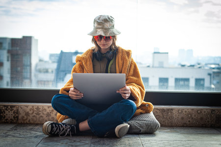 Hipster teenage girl sitting with laptop on terrace with view on city. Young happy woman in hat using laptop, shopping or working online from home outdoors. Freelance and freelancer.の写真素材