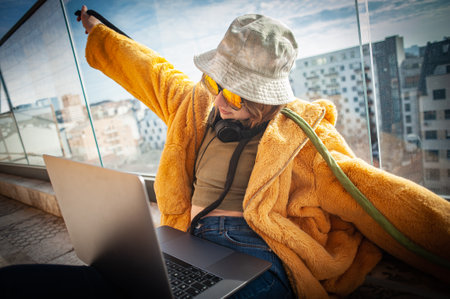 Copy Space. Hipster teenage girl sitting on terrace, cheerfully spreading her arms, using a laptop on her lap, with view on city and sky. Young happy woman in hat, shopping or working online from home outdoors. Freelance and freelancer.の写真素材
