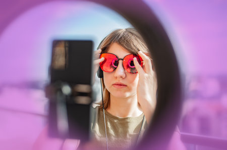 Teenage girl social media blogger recording video speaking looking at smartphone on tripod with ring light, on the balcony of the building.の写真素材