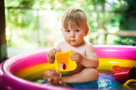 A cute little baby boy is playing with toys while bathing in a little colorful wading pool. Summer...
