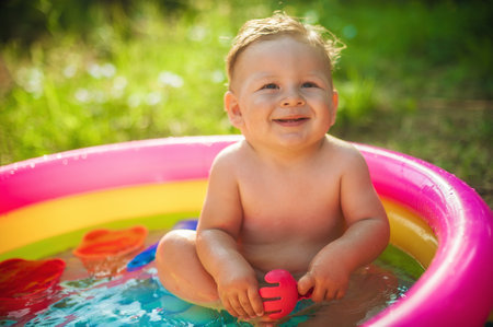 A cute little baby boy is playing with toys while bathing in a little colorful wading pool on meadow garden field outdoor. Summer time. Joy and happiness. Love and familyの写真素材