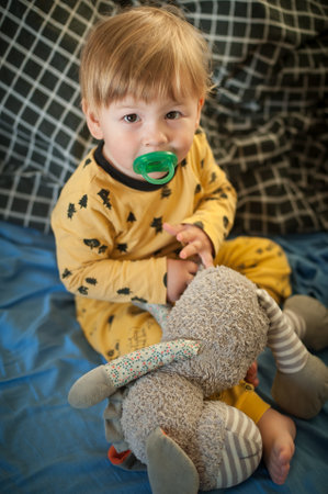 A toddler, wearing yellow pajamas with a black pattern, sits on a blue blanket. They are holding a soft, gray stuffed animal with striped legs and a floral pattern on one wingの写真素材