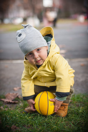 A young child wearing a cozy yellow raincoat and a playful hat leans over a bright basketball on the...