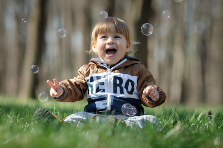 A smiling toddler sits on lush green grass in a tranquil forest, surrounded by floating bubbles that...