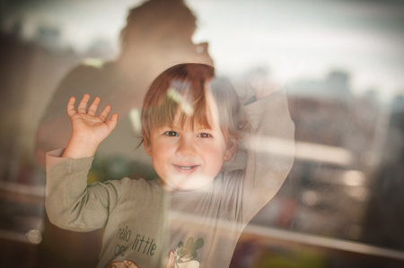 A young child enjoys a playful moment by the window, waving with excitement. Sunlight filters through, illuminating the cheerful expression on their face.の写真素材