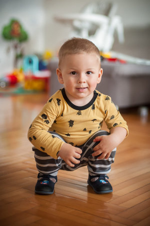 A cheerful toddler in a patterned long-sleeve shirt and striped pants squats playfully on a wooden...