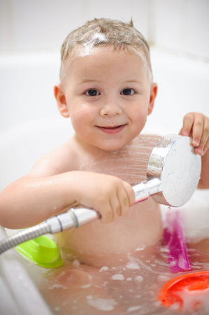 A cheerful toddler sits in a bathtub filled with bubbles and colorful toys, happily holding a...