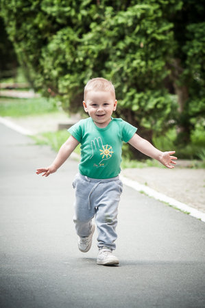 A young child in a green shirt runs energetically down a pathway in a park, smiling broadly and enjoying a sunny day amidst lush greenery.の写真素材