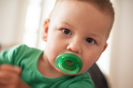 A toddler in a green shirt holds a pacifier while looking curiously at the camera. Soft light fills...