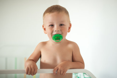 A young boy stands beside a crib, smiling and holding a green pacifier in his mouth. The room is bright and inviting, creating a joyful atmosphere.の写真素材