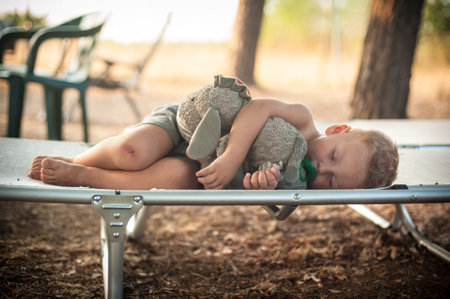 A child lies comfortably on a cot, holding a stuffed animal close while enjoying a peaceful nap in a tranquil outdoor environment. The warm afternoon light adds to the tranquil atmosphere.の写真素材