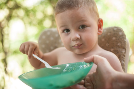 A young child sitting in a high chair is eating from a green bowl, showing curiosity and joy while being fed outdoors. The background features lush greenery, creating a cheerful atmosphere.の写真素材