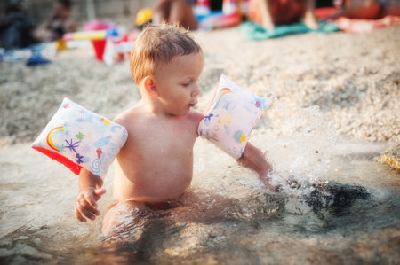 A young child sits in shallow water at the beach, wearing colorful arm floaties. The child splashes water joyfully, surrounded by bright beach toys and cheerful summer crowds.の写真素材