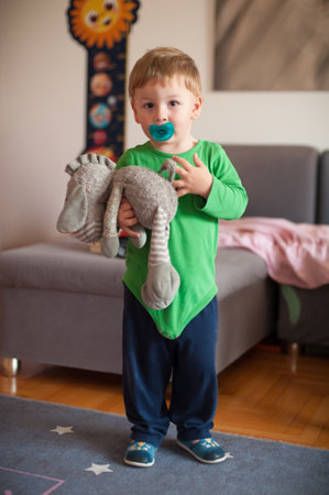 A young boy stands in a cozy living room, holding a stuffed animal close while sucking on a pacifier. He wears a green long-sleeve shirt and dark pants, smiling softly.の写真素材