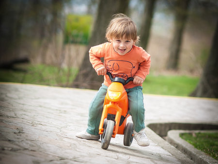 A young boy is joyfully riding an orange tricycle in a park, wearing an orange shirt and green pants. Lush greenery surrounds him, creating a cheerful atmosphere on a sunny day.の写真素材