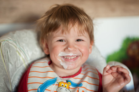 A toddler sits happily in a high chair, wearing a bib and smiling with yogurt on his face. The warm morning light highlights his cheerful expression in a cozy kitchen atmosphere.の写真素材