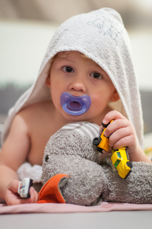 A young child relaxes under a towel, holding toy cars and a stuffed animal while using a pacifier....