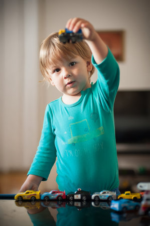 A young child wearing a blue shirt plays with a variety of toy cars on a table in a warm, inviting living room. The child holds a blue car high, showing excitement.の写真素材