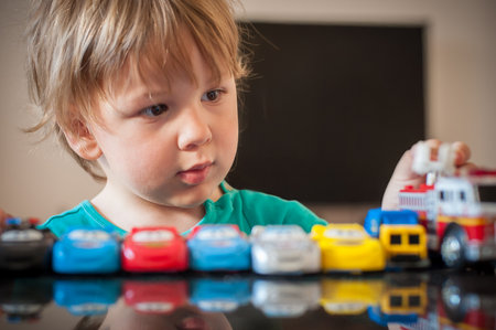 A little boy carefully lines up colorful toy cars and a fire truck on a table. His focused expression shows he is deeply engaged in imaginative play in a modern room.の写真素材