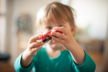 A child with light brown hair holds a red toy car close to their face, smiling while sitting in a...
