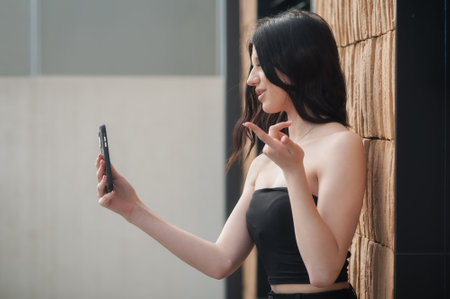 A fashionable young girl is taking a selfie in a stylish outfit against a contemporary textured wall. She strikes a playful pose, capturing the essence of social media influence during the day.の写真素材