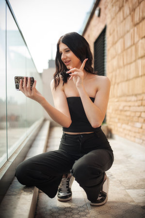 A fashionable young girl kneels on a stylish balcony, taking a selfie with her phone. She exudes confidence and modernity with her trendy outfit and playful attitude in the sunlight.の写真素材