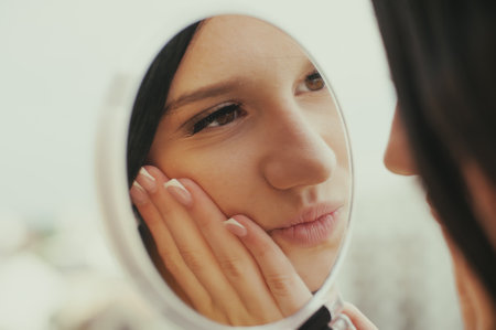 A young woman with black hair gazes into a hand mirror, touching her face. She is possibly checking her appearance or posing for social media, displaying a fashionable look.の写真素材