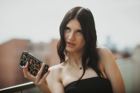 A stylish woman holds a phone while sitting on a balcony. She gazes thoughtfully into the distance, surrounded by a vibrant city backdrop under clear skies.の写真素材