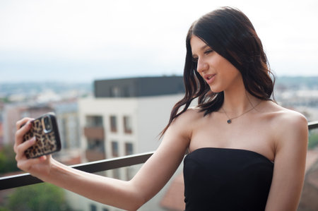 A young Woman is taking a selfie outdoors on a balcony wearing a black dress. She holds a mobile phone and is smiling slightly with an urban background.の写真素材