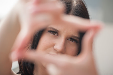 A young woman looks directly at the viewer. She uses her hands to create a rectangular frame around her face. The natural light illuminates her face and hands.の写真素材