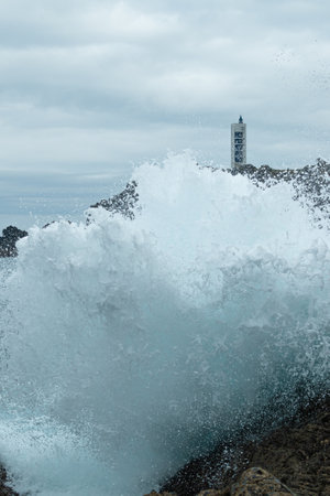 In the photo you can see a wave breaking against the cliffs and forming a great curtain of foam, in the background is the Frouxeira lighthouse, on the Galician coast.の写真素材