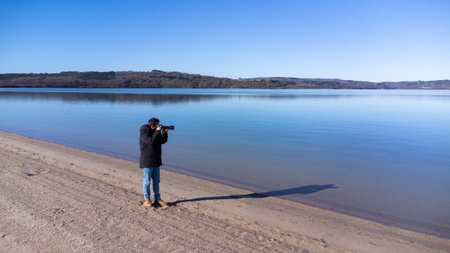 In the photograph a photographer is seen taking photos of a beach landscape with a blue sky and a calm lake.の写真素材
