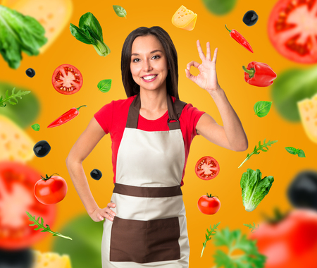 Cooking and food concept - smiling female chef, cook or baker with fork showing ok sign over falling vegetables on yellow backgroundの写真素材
