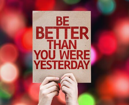 Hands holding cardboard with Be Better Than You Were Yesterday on bokeh backgroundの写真素材