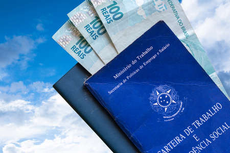 Brazilian Work Card with Brazilian money isolated on blue sky and clouds background. Written in Portuguese Federative Republic of Brazil, Ministry of Labor. Work and Social Security card. Brazilian work conceptの写真素材