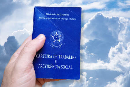 Man's hand holding Brazilian Work Card isolated on blue sky and clouds background. Written in Portuguese Federative Republic of Brazil, Ministry of Labor. Work and Social Security card. Brazilian work conceptの写真素材