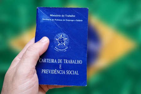 Man's hand holding Brazilian Work Card isolated on brazilian flag defocused background. Written in Portuguese Federative Republic of Brazil, Ministry of Labor. Work and Social Security card. Brazilian work conceptの写真素材