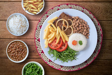 Brazilian food dish. Beans, rice, meat, chips, cabbage, tomatoes, onions. Executive dish. Dish done. Top view. Isolated on wooden backgroundの写真素材